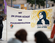 Zwei Personen halten ein Transparent, an dem Stöcke angebracht sind. Bild und Name von Jina Amini ist zu lesen, das Logo von TJK-E und "Mit Jin Jiyan Azadi zur Frauenrevolution" sind ebenfalls zu sehen.