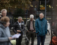 Im Vordergrund sind verschmonnene Menschen zu sehen, u.a. Irina Katz mit Blättern in der Hand vor einem Mikrofon. Im Hintergrund das Straßenschild "Gurs", im Fokus.