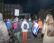 Eine Menschenmenge ist in Richtung einer Bühne auf dem Platz der Alten Synagoge gerichtet. Es ist dunkel. Vorne steht eine Person mit kleiner Israelfahne und langen, braunen Haaren. Etwas weiter hält ein Mensch ein Schild: "Free Gaza from Hamas".