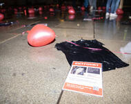 Ein schwarzes T-Shirt liegt auf dem Boden, darauf ein Steckbrief mit einer entführten Person auf nassem Boden. Daran angeheftet ist ein roter Herzchen-Luftballon. Es ist dunkel. Im Hintergrund: Beine und mehr Luftballons.