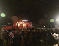 Auf dem Platz der Alten Synagoge blickt eine Menschenmenge auf eine Chanukkia. Es ist Dunkel. Von den Bildseiten strahlen zwei Lichter. Unten rechts steht Moshe Hayoun mit aufgeschlagenem Liedbuch.