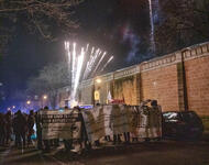 Eine Demonstration steht vor einer Gefängnismauer. Es ist Feuerwerk zu sehen und drei weiße Transparente.