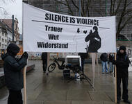 Zwei dunkel und winterlich gekleidete Menschen halten ein Banner mit zwei Holzlatten in die Höhe: Darauf abgebildet ein Polizist, der an einer schwarzen Person Gewalt ausübt. In schwarzen Druckbuchstaben steht: Silence is violence. Trauer, Wut, Widerstand