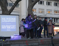 Menschen stehen auf dem Podest am Platz der Alten Synagoge, an einem Mikrofon. Links dahinter eine Leinwand, auf der ein Bild projeziert ist: Nie wieder ist Jetzt! Gegen jeden Antisemitismus! Solidarität mit Israel!