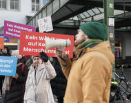 Ein Mensch mit Mütze und Schal spricht in ein Megafon. Dahinter sind Menschen mit Slogans auf farbigen Pappschildern aud enen steht "Kein WAhlkampf auf Kosten der Menschenrechte!" 