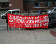 Demonstrierende auf einer Straße vor Hausfassade halten ein rotes Transpi auf dem steht: "Solidarität mit den 10 ehemaligen Militanten aus der RAF!"