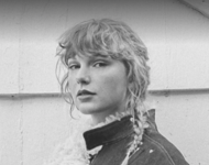 Die Sängerin Taylor Swift guckt auf einem Schwarz-Weiß-Foto in die Kamera. Sie trägt einen Wollmantel und hat einen geflochtenen Zopf, der ihr über die Schulter fällt. 