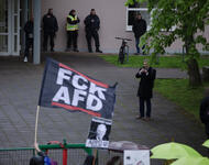 Im Vordergrund sind eine "FCK AFD"-Fahne zu sehen und ein Schild mit "Gauländer raus!", von der APPD und paar Schirme von den Omas gegen Rechts. Dahinter ist Robert Hagerman zu sehen, wie er den antifaschistischen Protest gegen die AfD abfilmt.