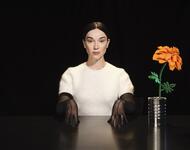 Die Sängerin St. Vincent schaut gerade in die Kamera, an einem schwarzen Tisch vor schwarzem Hintergrund sitzend, weiß gekleidet. Neben ihr steht eine orangene Blume auf dem Tisch.
