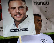 Armin Kurtović hält ein Schild mit dem Bild seines Sohnes Hamza Kurtović hoch. Er trägt ein weißes T-Shirt mit dem Konterfei seines ermordeten Sohnes. Im Hintergrund ist ein großes Banner zu sehen, ein Teil des Schriftzuges ist "Hanau".