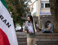 Am linken Bildrand eine Iran-Fahne mit Aufschrift "IRAN". Im Fokus eine männlich gelesene Person mit langen, schwarzen Haaren und Bart. Er blickt in Richtung der Fahne. Hält in den Händen ein Portrait von Toomaj Salehi und einen etwa 1,5 m hohen Galgen.