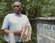 Eine Person steht neben einem Grabstein. Daran ist ein Holzkreuz gelehnt mit dem Namen Eliashukura Issack Nnko. Die Person trägt ein kariertes Hemd und hat einen Lappen in den Händen. Er schaut zur Seite, an der Kamera vorbei. Im Hintergrund stehen Bäume