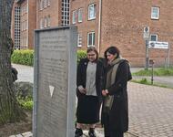Kelly Laubinger & Sahar Alias Baba Sheikh am Mahnmal der deportierten und ermordeten Sinti und Roma in Neumünster. Das Mahnmal - eine Platte, in die Namen der Opfer ausgestanzt sind- im Vordergrund; die zwei betrachten es. Im Hintergrund roter Klinker.