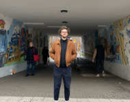 Niklas Schenker steht unter einer Unterführung mit Graffiti. Er trägt Turnschuhe und ein legeres Outfit mit Pulli und blauer Hose. Die Haare sind verwuschelt, die Hände in den Jackentaschen schaut er herausfordernd in die Kamera