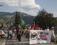 Auf einer schmalen Straße im Wohngebiet zieht eine Demonstration bergauf. Im Hintergrund Schwarzwald-Panorama, im Vordergrund rote und eine schwarz-grüne Fahne und Fronttransparent: "Es gibt kein ruhiges Hinterland" mit dem Logo der aufrufenden Gruppe.
