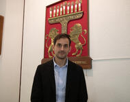 Phillip Peyman Engel (dunkle, kurze Haare, Schmales Gesicht, offenes, schwarzes Sakko auf hellblauem Hemd) steht vor dem Wappen der Israelitischen Gemeinde Freiburg (goldene Löwen vor einer Channukia auf rotem Grund) in der Neuen Synagoge.