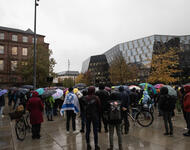Menschen mit Schirmen und Regenjacken stehen mit dem Rücken zur Kamera auf dem Platz der Alten Synagoge. Im Hintergrund rechts ist die Uni-Bibliothek und links das KG1 der Uni zu sehen. Eine Person hat eine Israel-Fahne über den Rücken gehängt.
