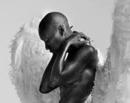 Cynthia Erivo ist im Bild fast nackt und hat zwei sehr große weiße, flauschig aussehendeFlügel auf dem Rücken. Ihr Kopf ist wie immer kahl rasiert und sie berührt im Bild mit beiden Händen ihren Rücken ziemlich direkt unterhalb ihres Nackens. 
