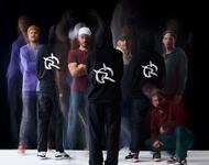 Irie Révoltés Pressefoto 2025 (c) Jamila K. Grote_2: Die neunköpfige Band teilweise mit schwarzen Kapuzenpullis und ihrem eigenen Logo vor dunklem Hintergrund. Einige Personen sind nur schemenhaft zu erkennen wie auf einem verwackelten Foto