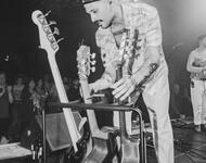 Ein Mann mit Kappe steht auf einer Bühne und greift zu einer Gitarre. Schwarz-weiß Foto.
