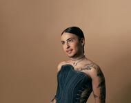 Kerosin85 schaut seitlich an der Kamera vorbei und lächelt ein wenig ironisch. They trägt ein schulterloses Jeansoutfit. Auf seinen Schultern und Armen sind Tattoos zu sehen. Der Hintergrund ist zwischen sand- und ockerfarben