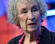 Die Autorin Margaret Atwood im Jahr 2022: Sie trägt die kurzen, weißen Haare gewellt; eine Kette mit großen pinken Perlen um den Hals, ein schwarzes Sakko und ein Mikrofon, das an einem Draht von hinter ihrem rechten Ohr vor zum Mund führt.