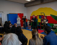 In einem Raum mit bunten Wänden stehen Menschen und schauen auf das Publikum. das mit dem Rücken zur Kamera links-unten im Bild zu sehen ist. Rechts sitzt Martin Horn. In der Bildmitte ist Sylvia Schliebe zu sehen, weitere Personen der Kita und Kinder.