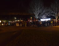Die Gegendemonstration mit mehreren Bannern und fahnen in einem eingezeichneten Bereich im Dunkeln mit Lcihtern, während diese zusammen singen.
