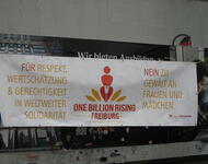 1 Banner zwischen 2 Bäumen. mittig steht rot auf weiß ONE BILLION RISING FREIBURG + Logo UND URL. Rechts und links steht in gelb: Für Restpekt, Wertschätzung & Gerechtigkeit in weltweiter Solidarität. Nein zu Gewalt an Frauen und Mädchen. Frauenhorizonte