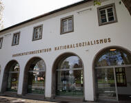 Blick von vorne auf das Gebäude mit Schriftzug "Dokumentationszentrum Nationalsozialismus". Unten fünf große gebogene Fenster. Oben kleine, viereckige Fenster. Links ist ein Teil einer Baumkrone mit frischen Trieben zu sehen.