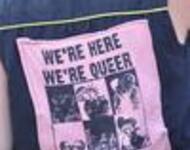 Ein Shirt auf dem ein Patch genäht ist. Darauf steht: We´re here, we´re queer, we riot 