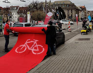 Roter Teppich mit Radwegzeichen wird über ein Auto gerollt, das auf dem Radweg parkt