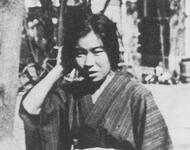 Kaneko Fumiko: Die junge Frau trägt einen Kimono und einen Gürtel. Das Schwarz-Weiß Foto ist im Freien aufgenommen. Sie hat eine Hand zum Kopf erhoben und schaut zur Seite auf den Boden. Im Hintergrund ist ein Fahrrad, ein Baumstamm & 1 Baracke erkennbar.