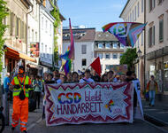 Demofront mit Transparent "CSD bleibt Handarbeit". Links ein neon-orange gekleideter Mensch mit Hundemaske. Im Hintergrund Gebäude der Emmendinger Innenstadt.